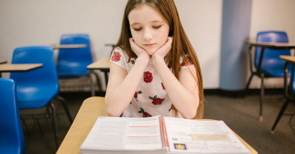 A young girl sits thoughtfully at her desk in a classroom, reading a textbook.